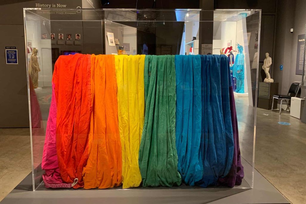 A view of a museum display case with the gathered folds of a piece of rainbow-striped cloth, including 8 bright colors from pink to purple