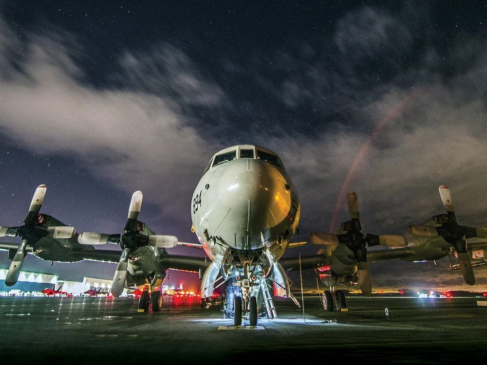 A massive aircraft with four propellers sits on a wet tarmac reflecting the bright red and white lights of the surrounding air base. The night sky above is cloudy, though some bright stars are visible.