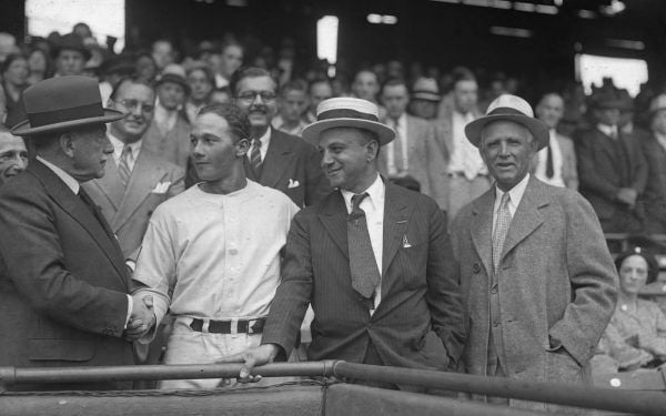 Joe Cambria stands in the foreground, smiling, flanked by onlookers in the stands and a baseball player