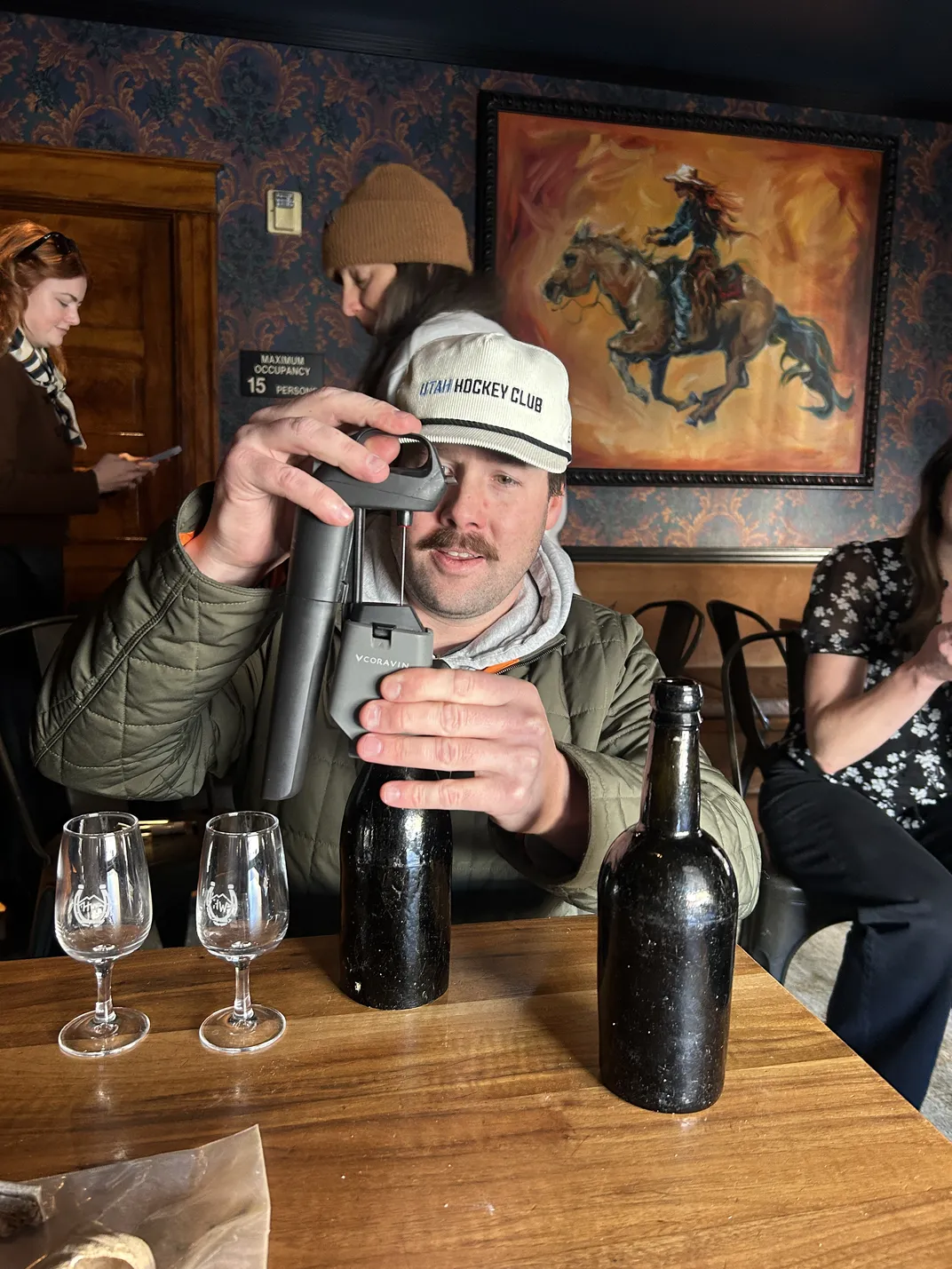 A man in a hat holding a bottle next to some glasses on a counter