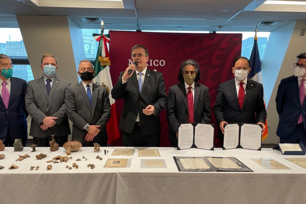 Seven people in suits and ties stand in front of a table with antiquities and 16th century documents laid out