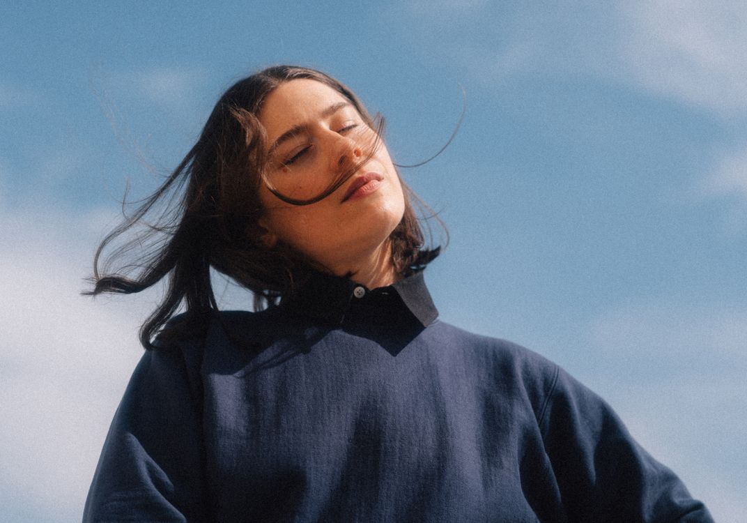 Person with blurred face stands under a blue sky with scattered clouds, wearing a dark blue sweater.