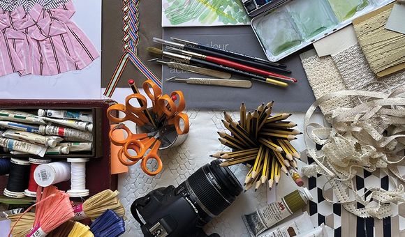 A variety of tools used to create art are displayed including scissors, fabric, a digital camera, tubes of paint and paintbrushes