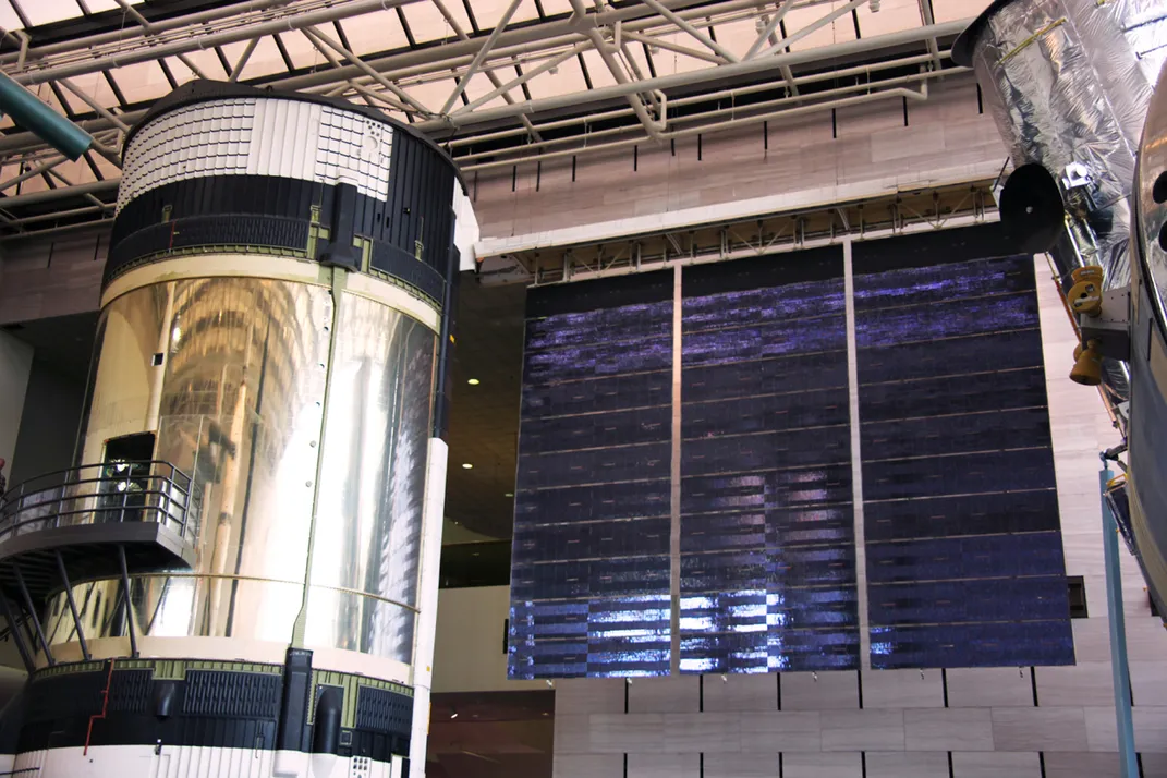 A cylindrical space station with a door and stairs leading up to it is on display in a museum with a wing of solar panels extending out