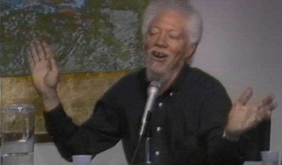 Benny Andrews sits at a table with a microphone, cups, and a pitcher of water with a yellow, green, and blue abstract painting hanging behind him. He is wearing a black button down shirt and raising his hands while talking.
