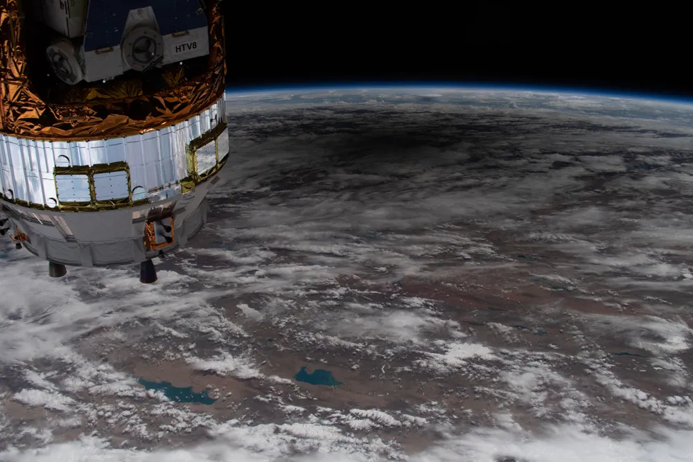 overhead shot of the earth with many clouds visible and a dark spot near the top. part of the ISS is seen at the upper left
