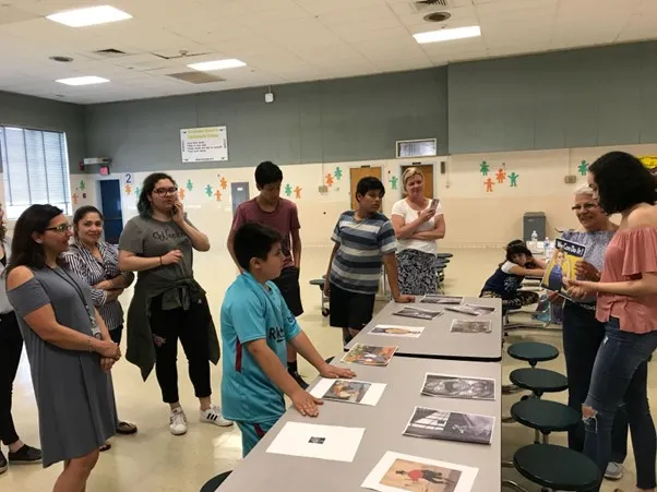 Group of adults and middle school children standing and actively discussing a set pf printed pictures laid out on a central table.