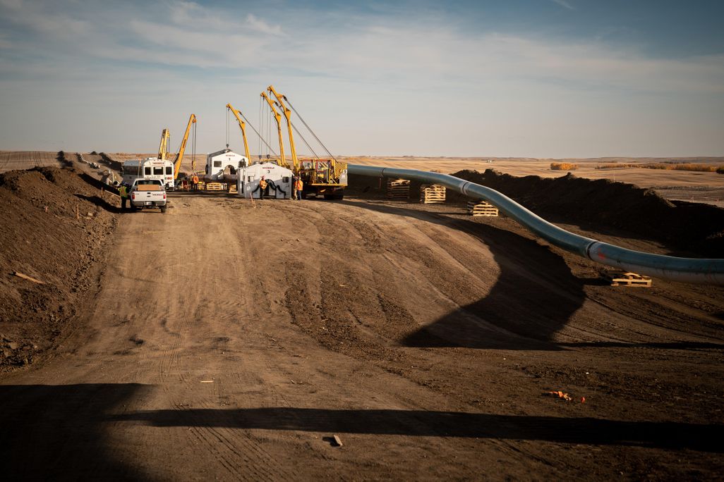 Five construction cranes, a truck and a bus sit atop a dirt road alongside pipeline, flanking the right side of the image.