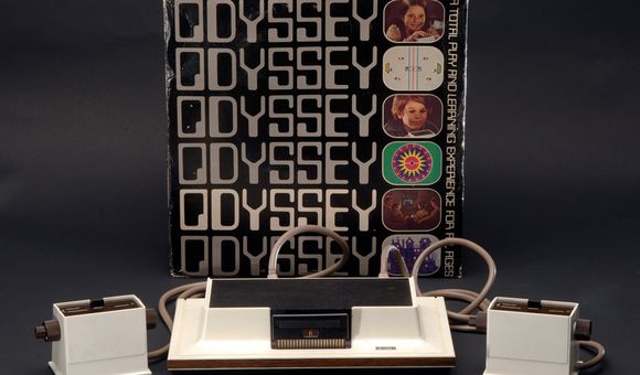 The Magnavox Odyssey system arrayed in front of its cover box. The box is decorated with the system's name and small images of various games and children using the Odyssey's controller
