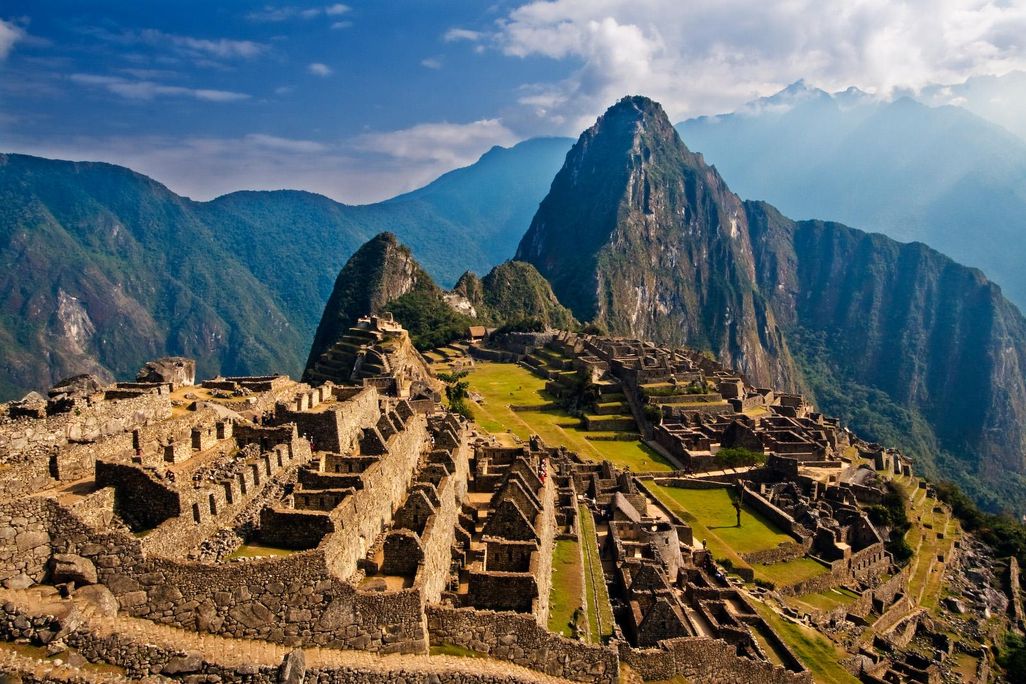 An aerial view of Machu Picchu at sunrise, with an orange light coloring the 15th century stone structures and a far off peak covered in green foliage