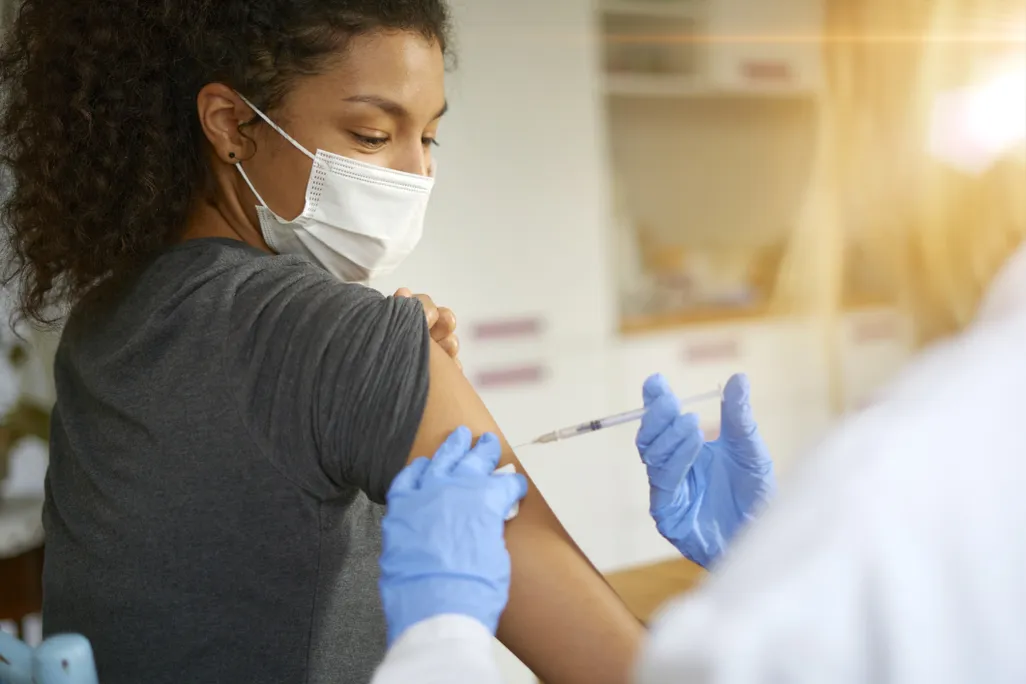 a woman in a mask gets a vaccine in her arm from a person in a lab coat and gloves