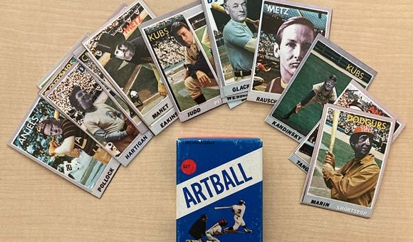 Baseball cards with faces of artists superimposed on them fanned out on a desk above a blue box with text that reads ARTBALL, an image of a batter, catcher, and umpire, with a red circle that says set 1.