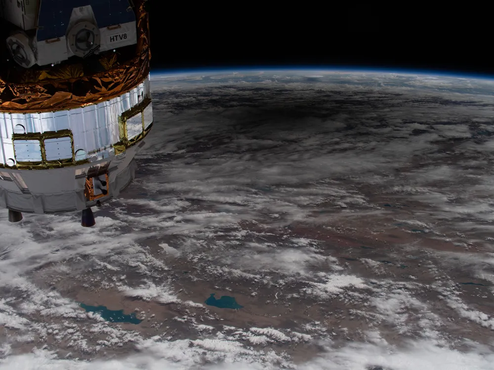 overhead shot of the earth with many clouds visible and a dark spot near the top. part of the ISS is seen at the upper left