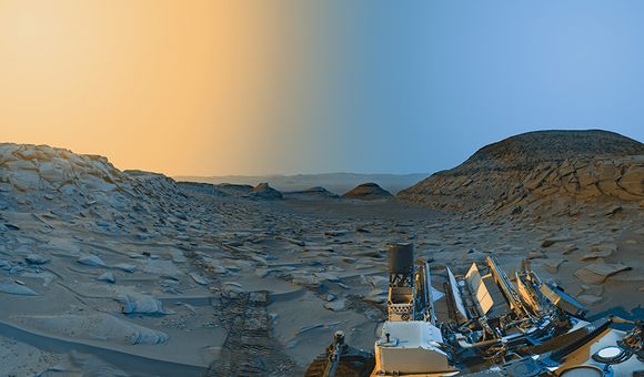 A stunning panorama of the Martian desert with a twilight sky. The landscape appears light rust and dull blue and is covered with boulders. A hill can be seen on the right.