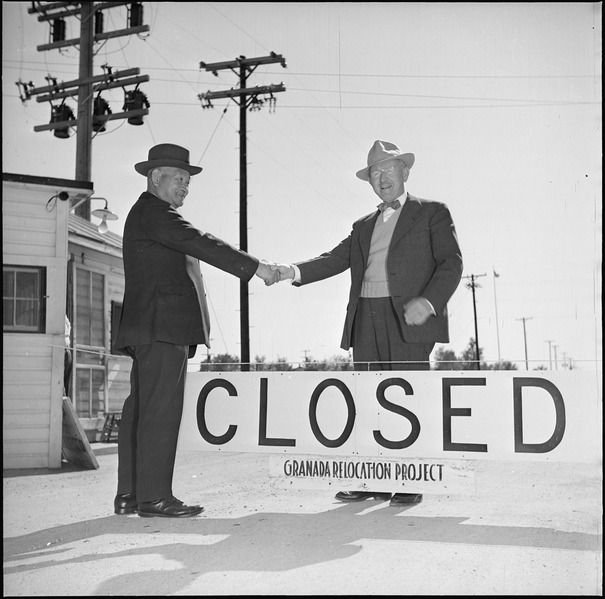 A black and white photo shows two men shaking hands in front of a sign that reads "Closed-Granada Relocation Project".