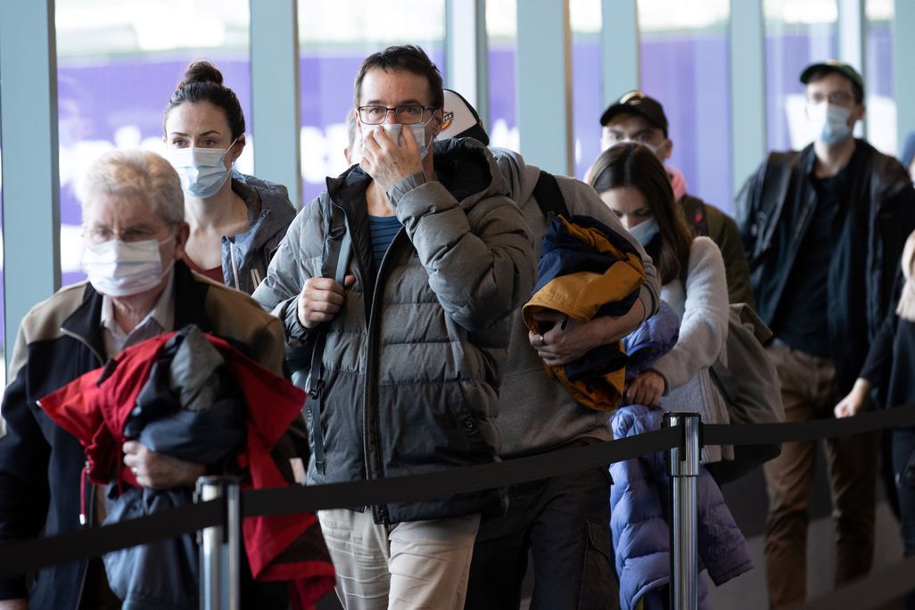 A crowd of people walk through an airport wearing masks. The man in the center of the image is touching his mask with one hand