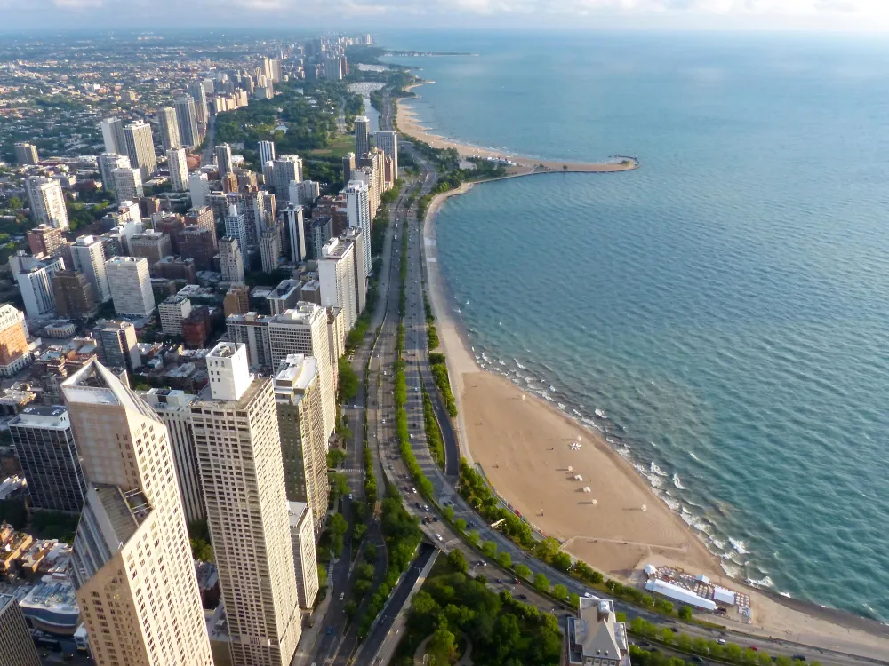 A bright aerial view on a sunny day of Chicago, with skyscrapers on the left and the curve of a beach and a major two-lane highway in the middle, and blue, calm Lake Michigan on the right