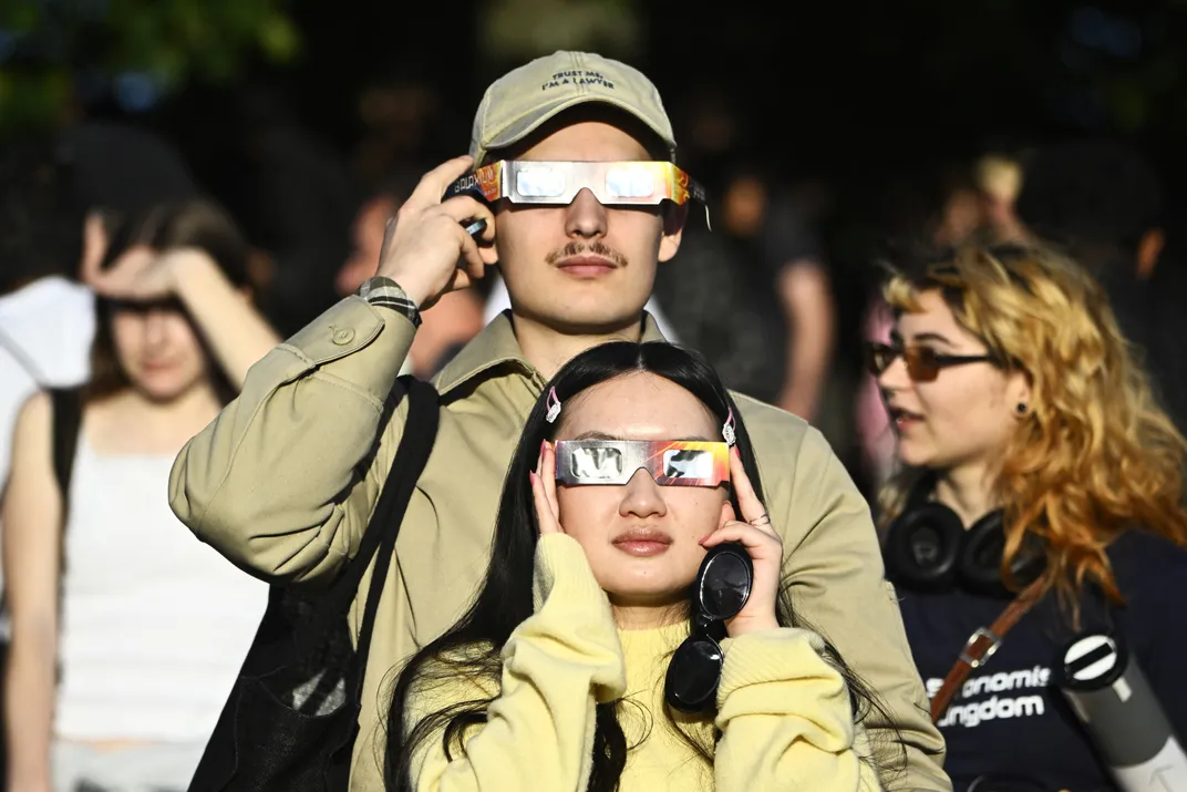 two people look toward the camera with eclipse glasses, the sun reflecting off
