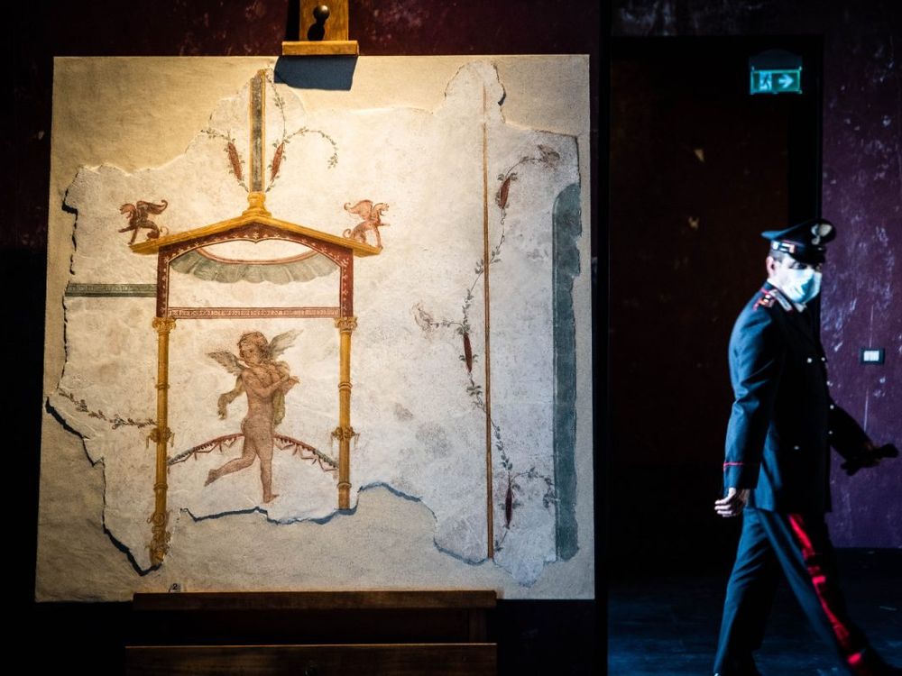 The recovered fresco fragment hangs on an easel display, lit up in foreground; in background, a police officer wearing a mask walks past the display