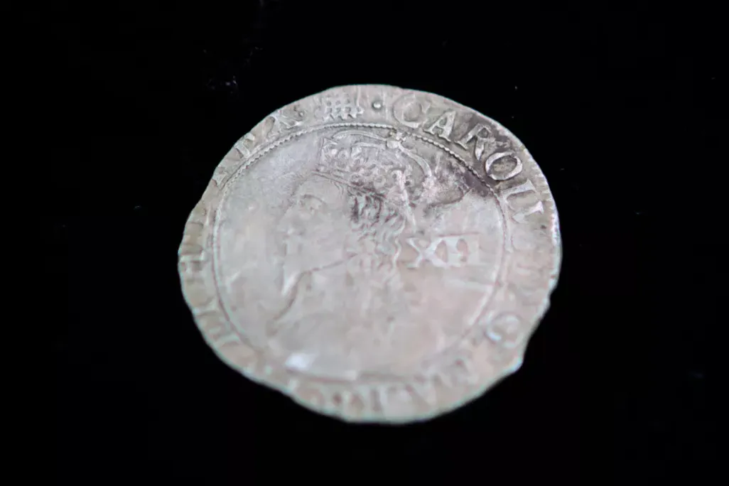 A silver coin with the face of a King and large capital letters going around in a circle; uneven edges
