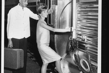 A black-and-white photograph of an African American male porter holding a suitcase while a white woman steps up into a rail car.