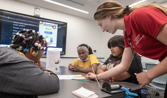 A young adult in a red polo points to a worksheet on a table. Three students are sitting at the table which has a table top rocket launcher, straw rocket, and table sign with instructions on top of it.