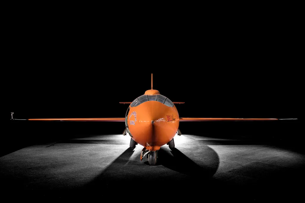 A bright orange plane, just big enough for one person to fit into, with cockpit facing the camera and each wing extended to either side, framed by dramatic lighting and a black background