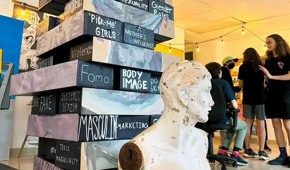 A pop-up museum display, featuring a tower of several “bricks” stacked like Jenga pieces. Each brick has writing on it: Fomo, body image, masculine marketing, pick-me girls, female sexuality, gender roles, a mother’s influence.