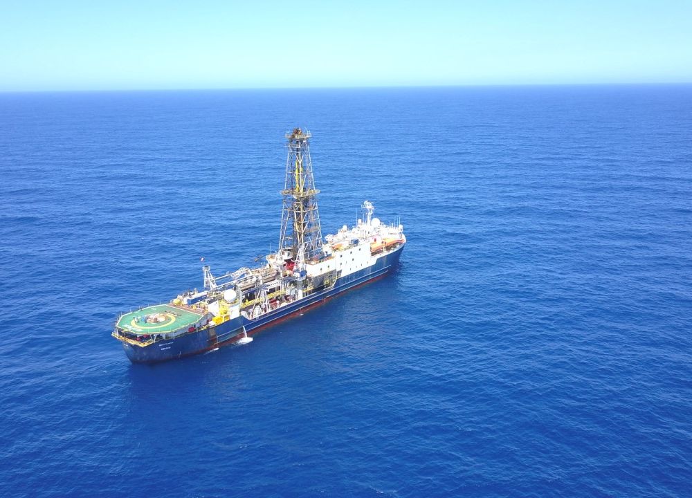A large research vessel with a cell tower on top sails across a dark blue stretch of ocean that expands towards a paler blue sky along the horizon.