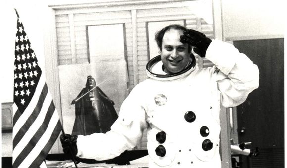 Informal classroom portrait of teacher Herbert Stephen Desind (1945-1992), wearing a reproduction Apollo-era spacesuit, holding an American flag; circa 1980s.  Desind was a space flight aficionado, and his collection of photographs of aircraft and spacecraft was donated to the National Air and Space Museum in 1997. This image is part of the Herbert Stephen Desind Collection.