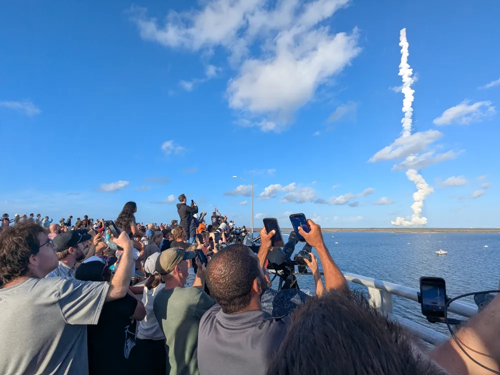people raise their phones to take pictures of the rocket launch, seen in the distance as a plume of white steam