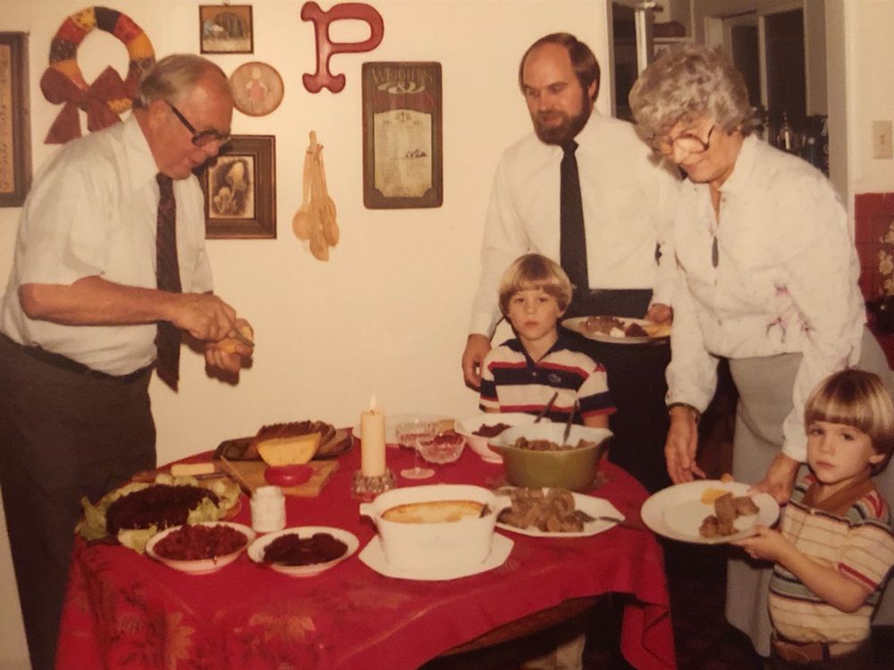 Three adults stand and two small children stand around a table with plated. The table is covered by a red table cloth and has a large candle in the center with food arranged around it.