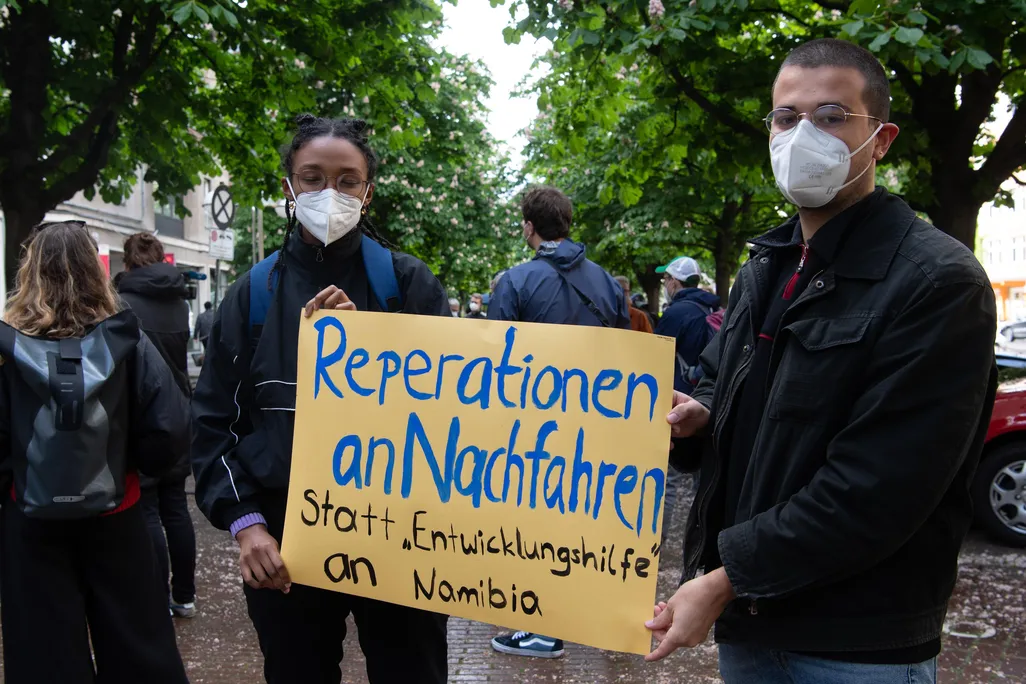 Two people in masks hold a yellow sign with blue and black handpainted letters, standing in a city street in front of trees and other protesters