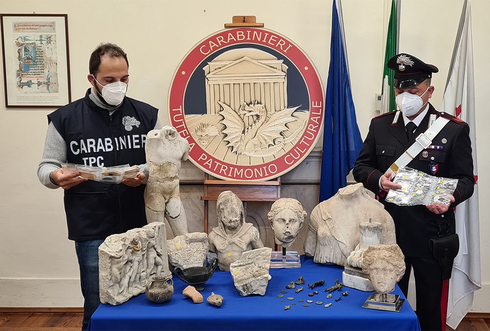 Two people in police uniforms stand behind a table covered in blue tablecloth, upon which sit several fragments of sculptures