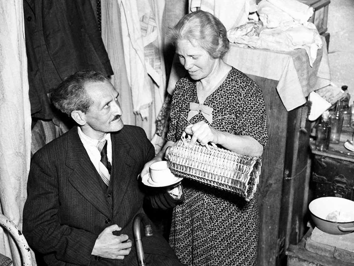 A white woman in a dress pours tea for a seated man in a suit, in a small home crowded with items (longform main)
