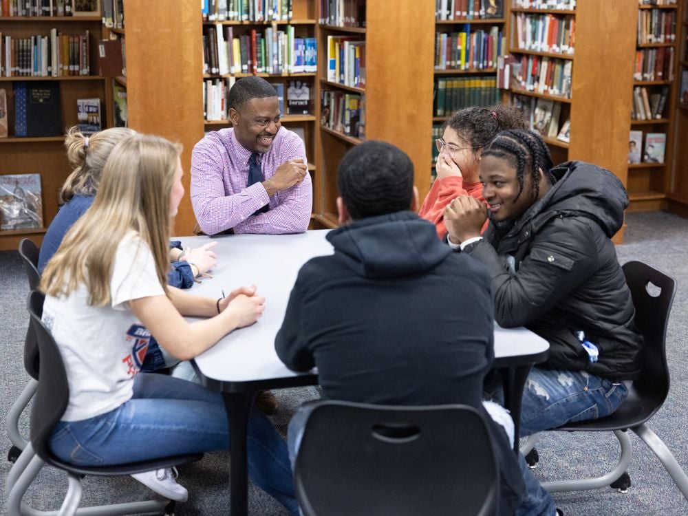 Teacher Kurt Russell sits at a square table in the school's library with five students, smiling and in conversation.