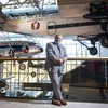 A Black man in his early 70s, wearing a blue suit and brown dress shoes, stands on a mezzanine overlooking a museum gallery displaying at least five airplanes.