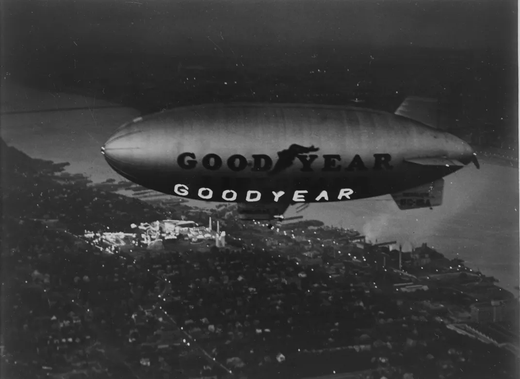 Black and white photo showing a Goodyear blimp with a lighted neon sign that reads GOODYEAR