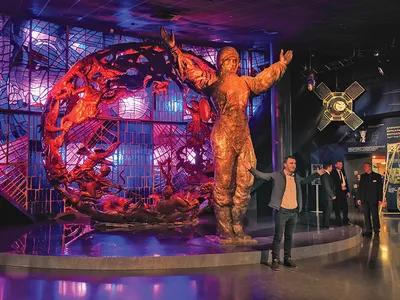 A museum display features a statue of a male astronaut standing with his arms outstretched in front of a metal sculpture lit by red and purple lighting. A museum visitor stands in a similar pose in front of the statue.