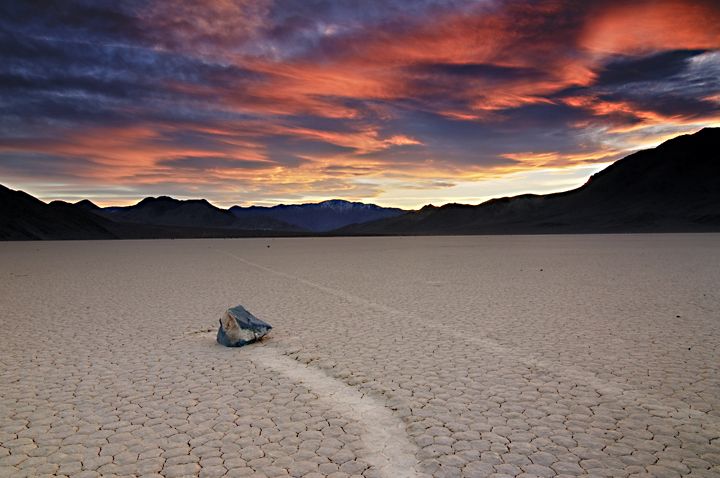 a rock with a trailing path is seen in desert terrain at sunset