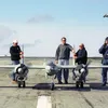 Four men stand on a paved runway behind a large, grey twin-engine remote-controlled model airplane. The man on the far right holds a radio transmitter with a long antenna, while a small helicopter is visible in the sky behind him. The scene is set under