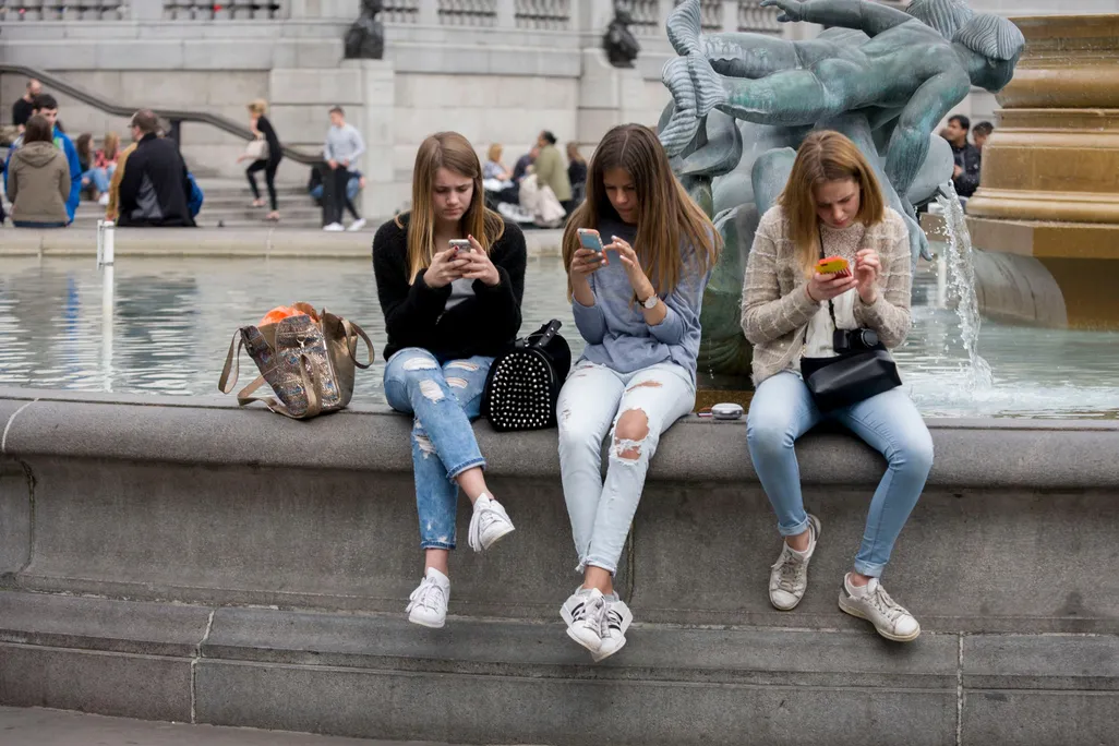 Three teenagers sit on the side of a large fountain and look at their smartphones