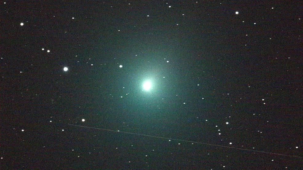 A photo of Comet 46P/Wirtanen. In the middle of the image is a ball of light that is glowing bright green against a starry black sky. 