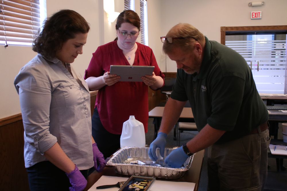 Two caucasian women stand at a table. A caucasian man leans over the table to hold wet cloth in an aluminum pan.