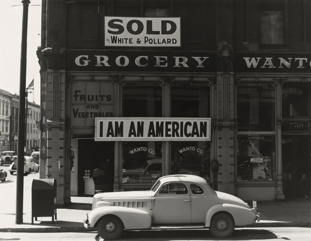 A black and white image of a grocery store front with a large sign in bold font: I AM AN AMERICAN