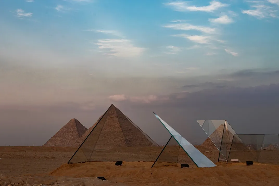 Huge shards of glass stick up from the sand at odd angles, in front of two large pyramids
