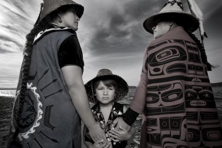 Darkfeather Ancheta, Eckos Chartraw-Ancheta, and Bibiana Ancheta, Tulalip, Washington. Darkfeather, is pictured with her sister Bibiana and nephew Eckos at the edge of Tulalip Bay. They are wearing traditional regalia prepared for their annual Canoe Journey. Every year, upwards of 100 U.S. tribes, Canadian First Nations and New Zealand canoe families will make “The Journey” by pulling their canoes to a host destination Tribe. Canoe families pull for weeks, and upon landing participate in several days and nights of “protocol”, a celebration sharing traditional knowledge, ancestral songs, and sacred dances. Photo by Matika Wilbur for Project 562. Courtesy of the Artist. 