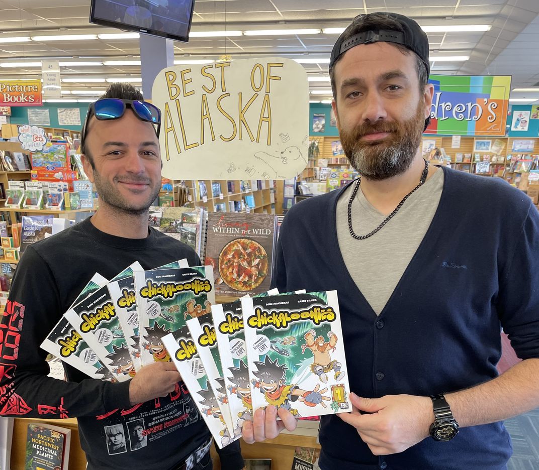 Casey Silver and Dimi Macheras stand in the foreground of a photograph holding copies of their comic book Chickaloonies: First Frost. They are posing at a book signing held at Title Wave Bookstore in Anchorage, Alaska.