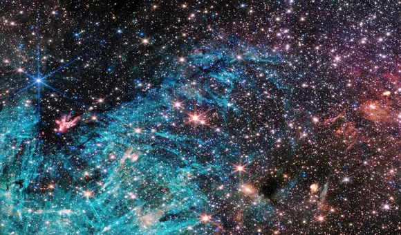 A multitude of stars packed together in a dense region of space, shining brightly in the surrounding darkness. A bright blue cloud in the lower half of the image with pitch black patches appearing within the cloud
