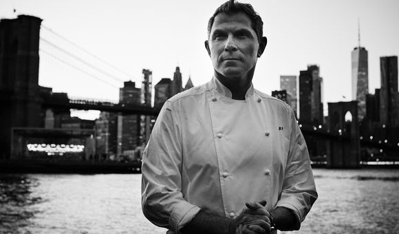 A black and white photograph of Bobby Flay in his signature white chef’s coat, with his initials, BF, embroidered on the front.